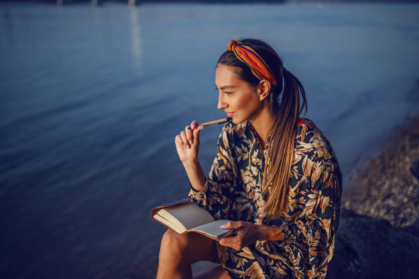 Portrét zamyšlené atraktivní bělošské brunetky v květinových šatech a s čelenkou sedící na skále na břehu u řeky a psacím deníkem. - Fotografie, Obrázek