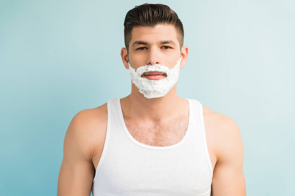Уверенный красивый молодой человек с кремом для бритья на лице с пустым выражением лица во время зрительного контакта в студии
 - Фото, изображение