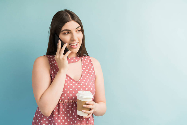 Schöne weibliche Millennial spricht auf dem Smartphone, während sie einen Drink vor einem schlichten Hintergrund hält - Foto, Bild