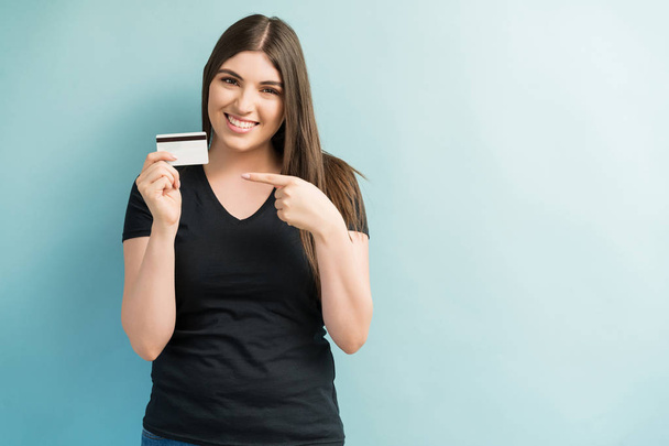 Attractive female smiling while pointing at credit card against plain background - Valokuva, kuva