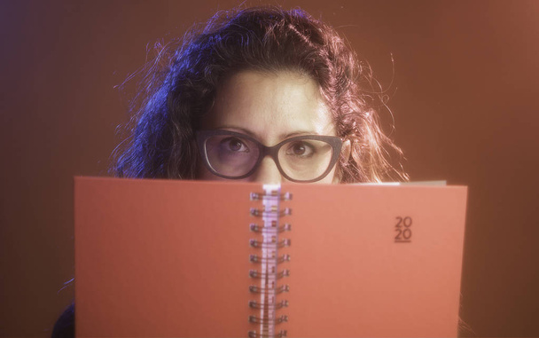 Femme d'âge moyen hispanique, tenant un agenda rouge de la nouvelle année 2020 et regardant la caméra. Utiliser des lunettes
. - Photo, image