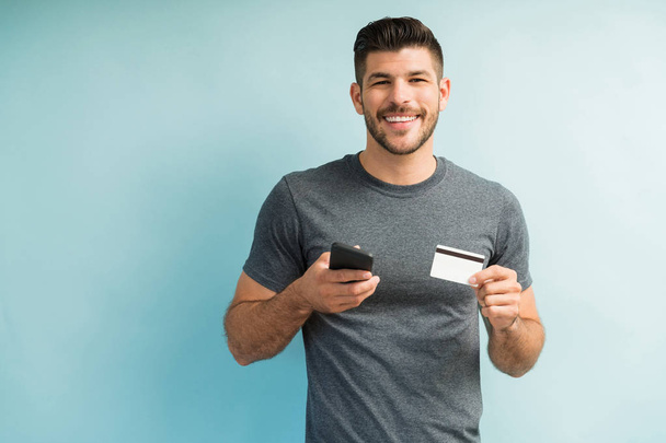Heureux jeune homme intelligent millénial tenant carte de crédit et téléphone mobile sur fond turquoise
 - Photo, image