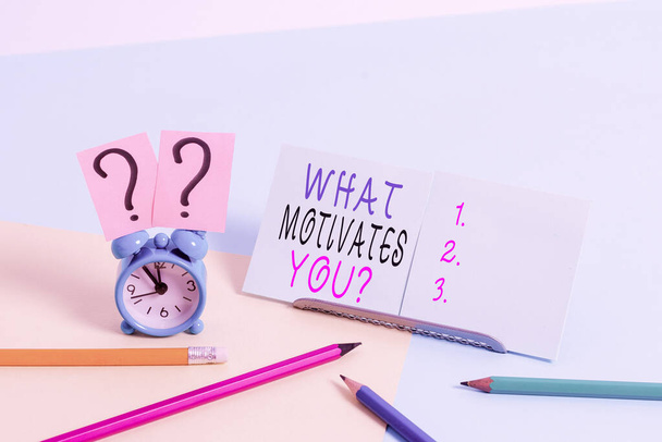 Writing note showing What Motivates Youquestion. Business photo showcasing know reasons why you want to wake up each morning Mini size alarm clock beside stationary on pastel backdrop. - Photo, Image