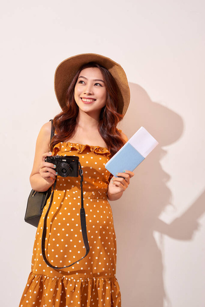 Πορτρέτο του μια ευτυχισμένη νεαρή γυναίκα με καπέλο κρατώντας κάμερα και δείχνει διαβατήριο ενώ στέκεται απομονωμένο πάνω μπεζ φόντο - Φωτογραφία, εικόνα