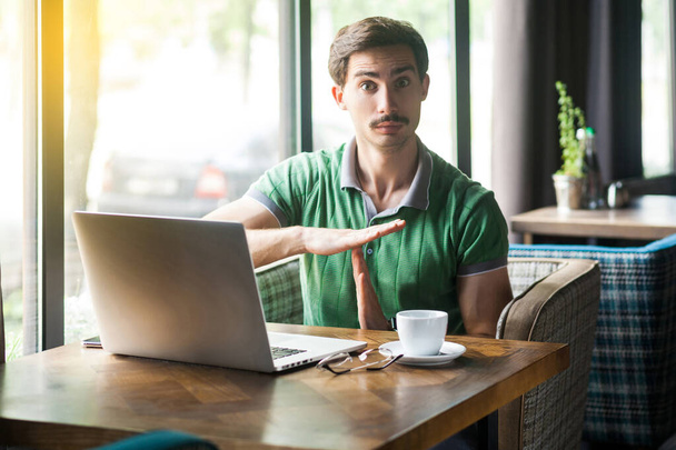 Young businessman in green t-shirt looking at camera and showing time gesture while sitting at table with laptop in cafe  - Photo, image