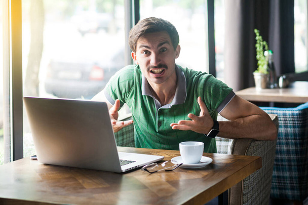 Young aggressive businessman in green t-shirt looking at camera with angry crazy face while sitting at table with laptop in cafe, business and freelancing concept - Fotoğraf, Görsel
