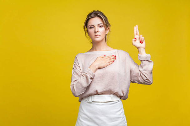  faithful serious woman with fair hair in casual blouse standing with hand on chest and fingers up while taking oath on yellow background - Photo, Image