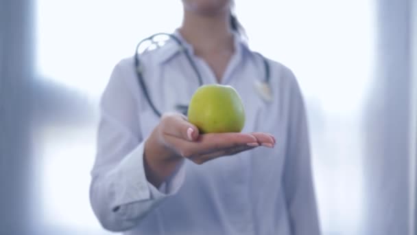Ellerinde yeşil elma olan doktor, beyaz arka planda sağlıklı beslenme tavsiyesi veren kameraya uzanıyor. - Video, Çekim