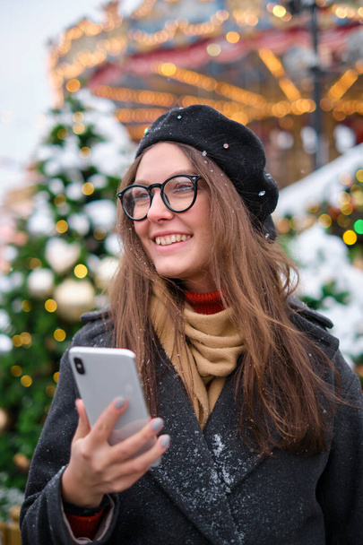young attractive girl uses a phone on the street, against the background of dressed-up Christmas trees, Christmas mood, - Foto, Imagen