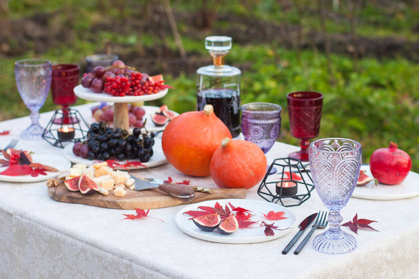 Outdoor standing decorated table with cheese platter on wooden board, pumpkins, grapes, figs, burning candles, colored glasses, bottle of wine and red autumn maple leaves. - Foto, Imagem