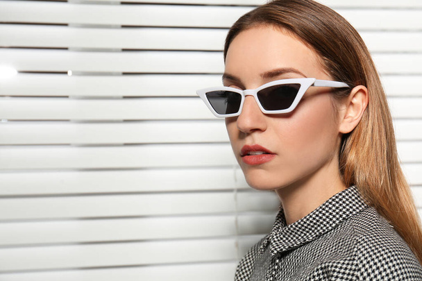 Young woman wearing stylish sunglasses against blinds - Photo, Image