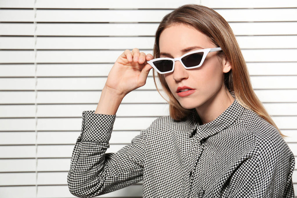 Young woman wearing stylish sunglasses against blinds - Фото, зображення
