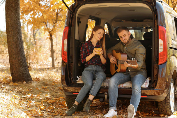 Young couple with guitar sitting in open car trunk outdoors - Foto, Bild