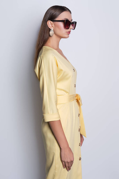 Fashion model in big sunglasses wearing yellow dress with buttons posing over gray background - Zdjęcie, obraz