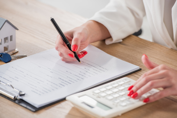 обрезанный вид предпринимательницы с помощью калькулятора и письменной форме в контракте рядом с моделью дома и ключи
 - Фото, изображение