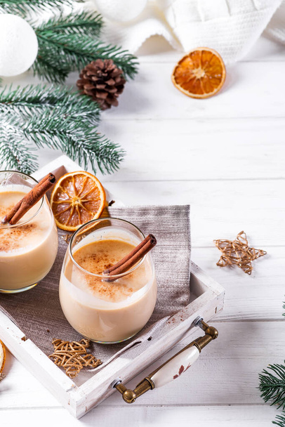 Winter spicy hot drink eggnog in a glasses with cinamon in tray, dry orange,fir branches and pine cone on white wooden table - 写真・画像
