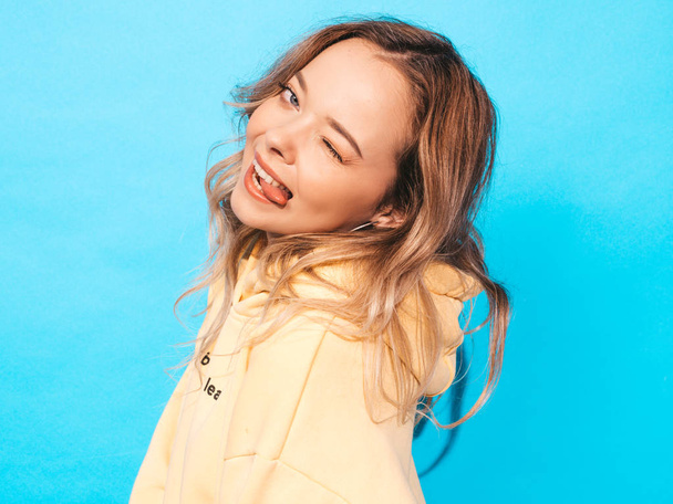 Portrait of young beautiful smiling girl in trendy summer hipster yellow hoodie.Sexy carefree woman posing near blue wall. Positive model having fun.Shows tongue and winks - Photo, image