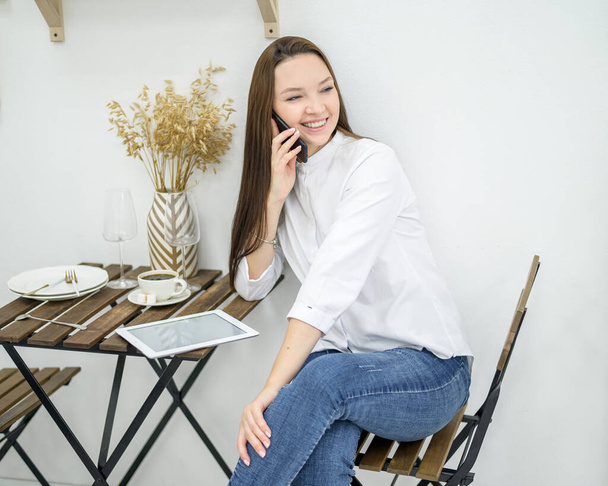 A woman in a white shirt and jeans sits in a cafe with a tablet . Female office worker at the lunch break talking on the phone. Manager at a business meeting in a restaurant drinks coffee. - Valokuva, kuva