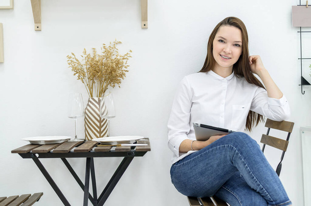 A young woman in a white shirt and jeans sits in a cafe at a table with a tablet in her hands. Female office worker at lunch break. Manager at a business meeting in a restaurant. - Foto, Bild