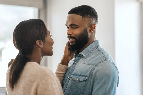 Young African American man smiling and looking at his wife while standing arm in arm together in their modern apartment - Photo, image