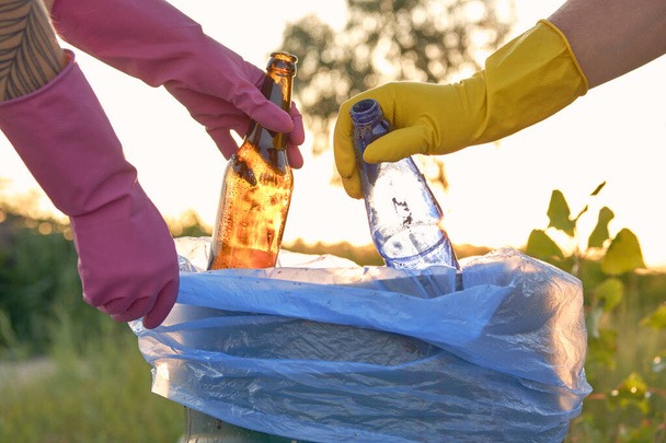Wolontariusze w fioletowo-żółtych rękawiczkach spacerują z workami na śmieci wzdłuż brudnej plaży rzeki i sprzątają śmieci. Zbliżenie. - Zdjęcie, obraz
