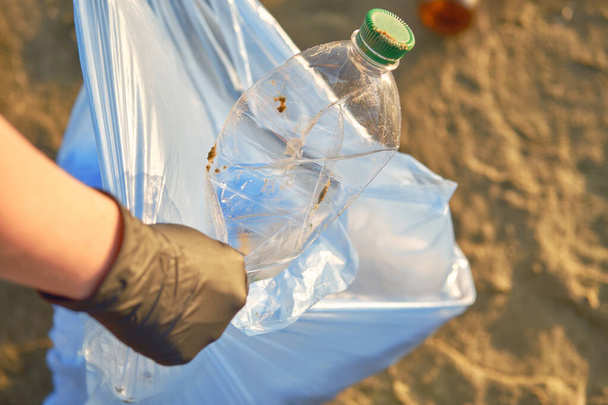Joven voluntario en guantes negros está caminando con bolsas de basura a lo largo de una playa sucia del río y limpiando basura. Gente y ecología. Cierre. - Foto, imagen