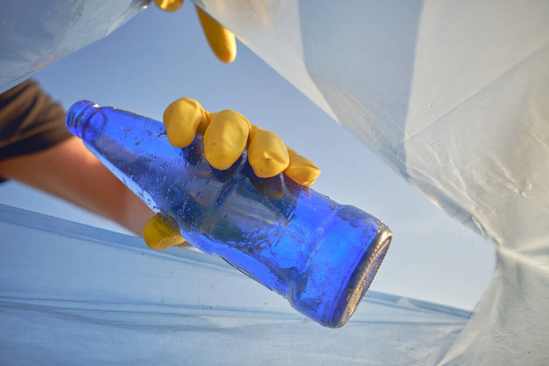 Hand einer bewussten Person in einem gelben Gummihandschuh legt eine gebrauchte blaue Glasflasche in einen Müllsack. Blauer Himmel. Uferverschmutzung. Nahaufnahme. - Foto, Bild