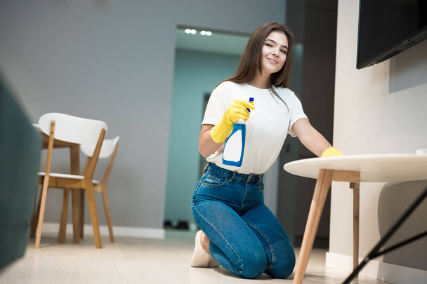cute young woman in yellow gloves with detergent spray in her hand wiping dust off from the kitchen chair with a rag - Photo, image