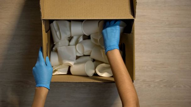 Person in protective gloves sorting paper cups in cardboard box, recycling - Φωτογραφία, εικόνα