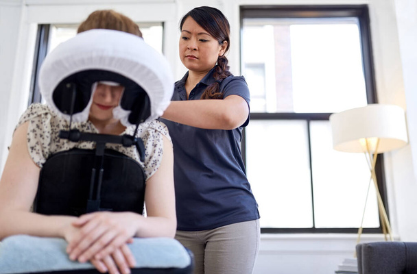 Chinesische Masseurin gibt einer attraktiven blonden Kundin an ihrem Arbeitsplatz in einem hellen Büro eine Nacken- und Rückendruckbehandlung - Foto, Bild