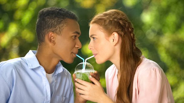 Multiracial couple drinking fresh cocktail through straws looking each other - Foto, imagen