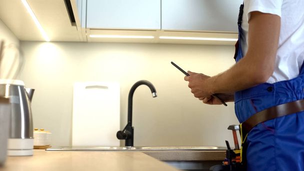 Handyman in uniform checking kitchen tap, typing report in tablet, plumbing - Foto, Imagem