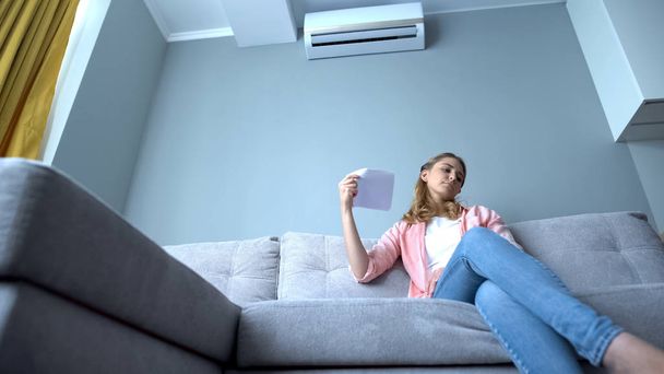 Woman waving paper sitting on couch, suffering heat, faulty air conditioning - Foto, afbeelding
