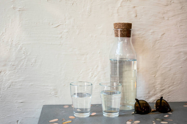 Pullo vedellä, kaksi lasia ja aurinkolasit pöydällä vasten valkoista seinää kahvilassa. Virkistävä kylmä vesi kuuman kesän auringon jälkeen
 - Valokuva, kuva