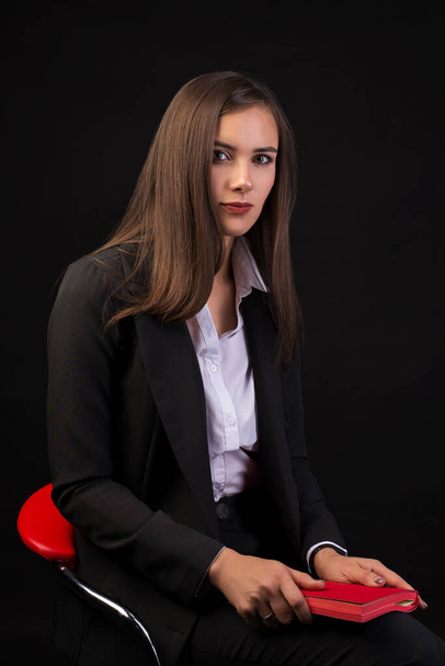 young beautiful girl with long black hair sitting on a red chair and holding a red notebook, black background, business suit girl - Фото, изображение