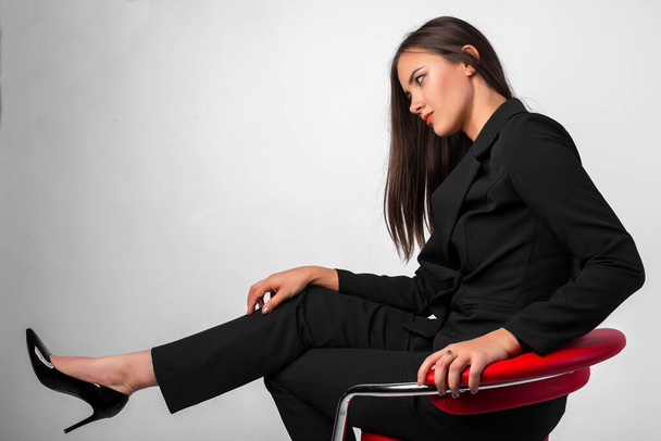 Young beautiful girl in business suit black jacket, pants, shoes sitting on red chair, white background, business lady, young cute brunette - Foto, Bild