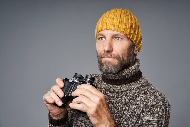 Focused mature man in warm sweater and knitted hat holding binoculars - Foto, Imagem