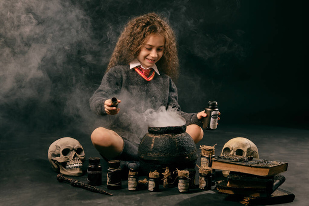 Little witch girl with long hair, dressed in dark clothes, is sitting near a steaming pot against a black studio background and preparing a potion. - Photo, Image