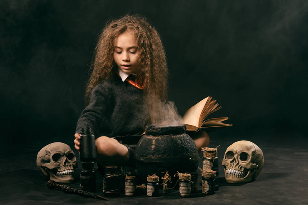 Klein heks meisje met lang haar, gekleed in donkere kleren, zit in de buurt van een stomende pot tegen een zwarte studio achtergrond en het bereiden van een drankje. - Foto, afbeelding