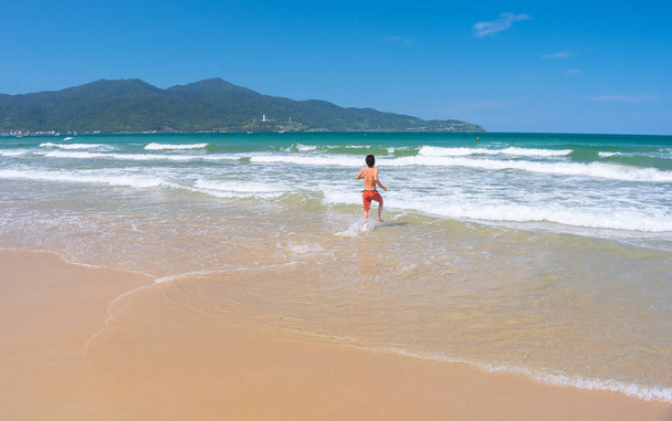 Da Nang Beach: an excited man runs through the surf to the gentle sea waves on My Khe Beach. Son Tra Peninsula and mountain on the horizon. This is the central beach of Danang City, Vietnam, in May. - 写真・画像