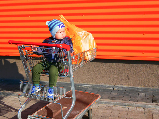 baby on a shopping trolley. Baby boy sitting in a shopping trolley outside. On orange background - Zdjęcie, obraz