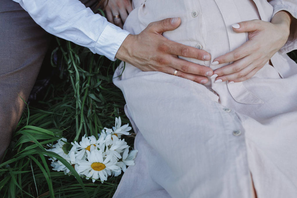 Crop of husband and pregnant wife, together sitting on green grass, holding by hands woman`s belly and expecting to baby. - Valokuva, kuva