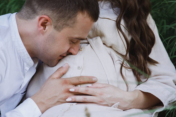 Урожай прекрасного мужа, трогающего и целующего ее беременную жену к животу. Красивая женщина сидит на траве рядом со своим мужчиной
. - Фото, изображение