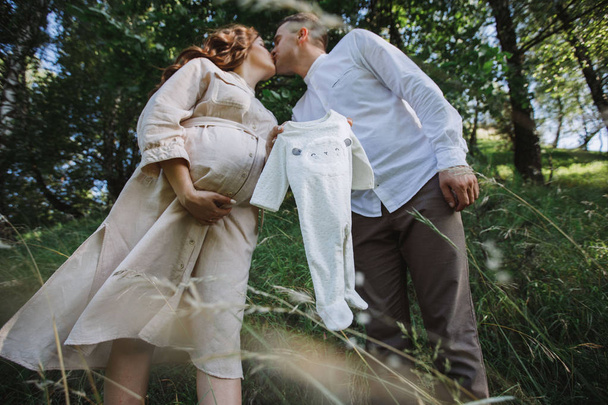 Wife and husband relaxing on wonderful nature, standing and kissing each other. Woman and man holding by hands their baby clothes. - Foto, afbeelding
