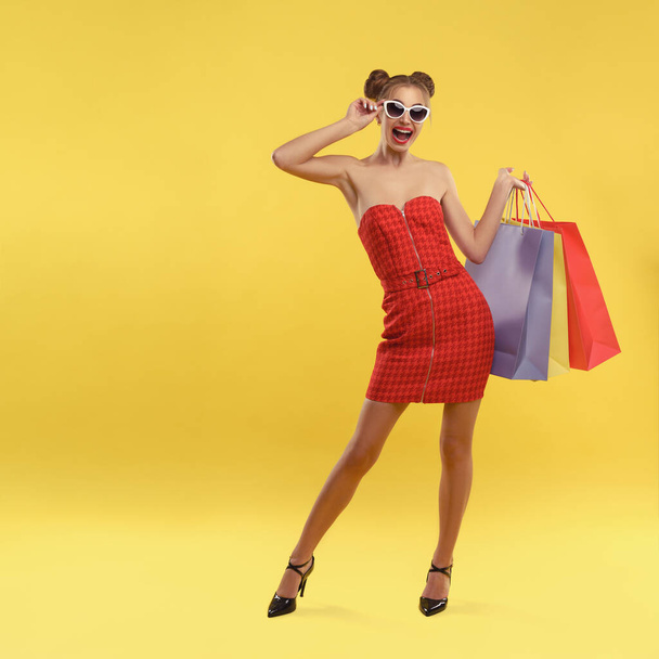 Щаслива дівчина в червоній сукні і сонцезахисних окулярах тримає барвисті сумки для покупок на жовтому тлі
 - Фото, зображення