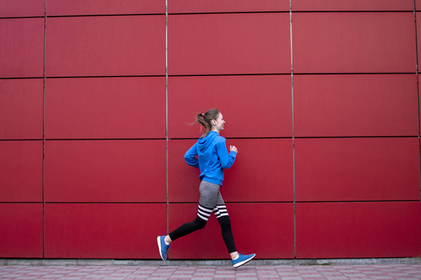 sporty girl runs against a red wall in the street, a woman jogging in sportswear outdoors, copy space - Photo, image