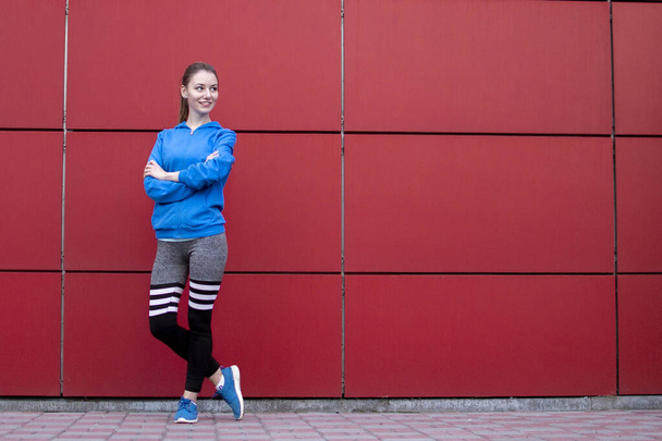 Спортивная девушка стоит напротив красной стены в спортивной одежде, фитнес-женщина позирует на городской улице, копировальное пространство
 - Фото, изображение