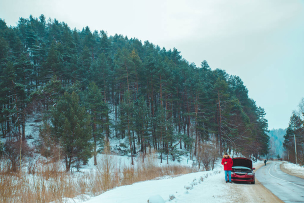Человек, стоящий рядом со сломанной машиной на обочине дороги в снежную зимнюю погоду
 - Фото, изображение