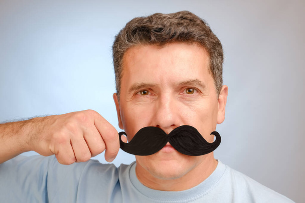 Portret mężczyzny ze sztucznymi wąsami w ręku, aby wziąć udział w listopadowym wydarzeniu mającym na celu pomoc mężczyznom w problemach zdrowotnych. - Zdjęcie, obraz