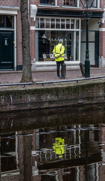 henkilö ja sen heijastus keltaisessa takissa olevasta henkilöstä kanavan vedessä näyteikkunan edessä
 - Valokuva, kuva